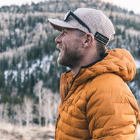 A man in profile wearing a tan mesh baseball cap with black sunglasses on the brim and a mustard yellow puffer jacket, set against a blurred mountain background with trees and snow.