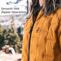 A close-up of a person wearing a mustard yellow quilted puffer jacket outdoors, with the text 'Smooth YKK Zipper Operation' in the upper left. The jacket has a small black embroidered bird logo on the chest and a black zipper running down the front.
