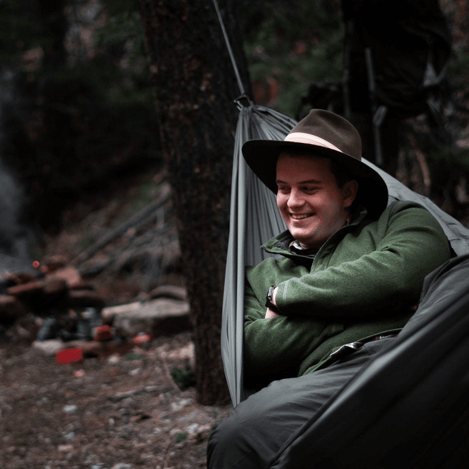 A camper wearing a brimmed hat lounging comfortably inside an Outdoor Vitals Ultralight Complete Hammock System at a wooded campsite.