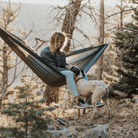 A person sitting in an Outdoor Vitals Ultralight Complete Hammock System while petting a dog outdoors in a forest.