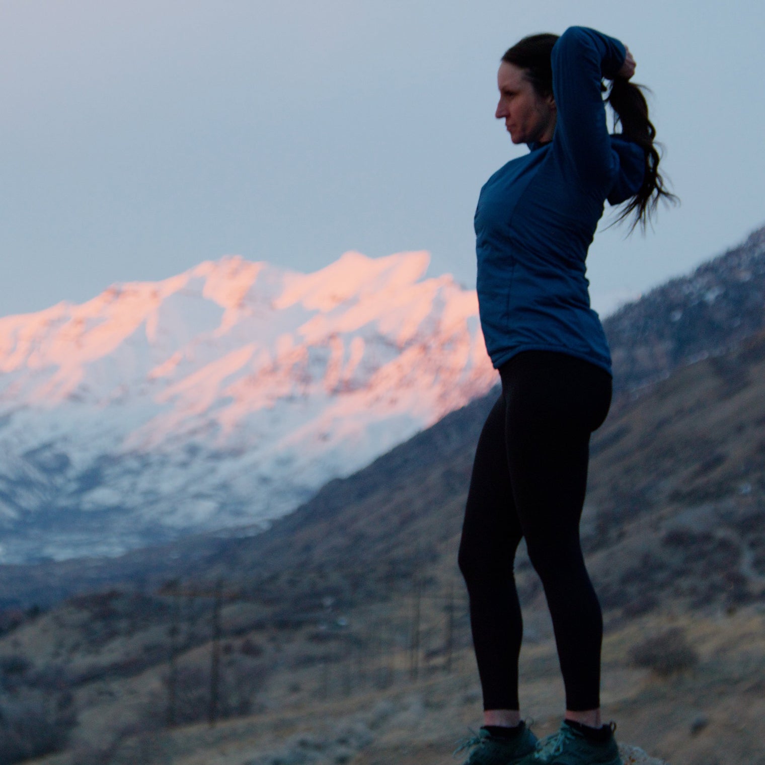 Side view of a woman tying her hair before a workout, wearing a Ventus Active Hoodie and leggings with snow-capped mountains behind her.