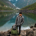 A man wearing a NovaUL down jacket stands by a scenic mountain lake next to a backpacking pack.