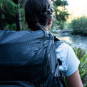 Close-up rear view of a person wearing the Outdoor Vitals KotaUL Ultralight Travel & Adventure Backpack, highlighting the durable grid-patterned ripstop fabric texture against a blurred outdoor forest and river backdrop.