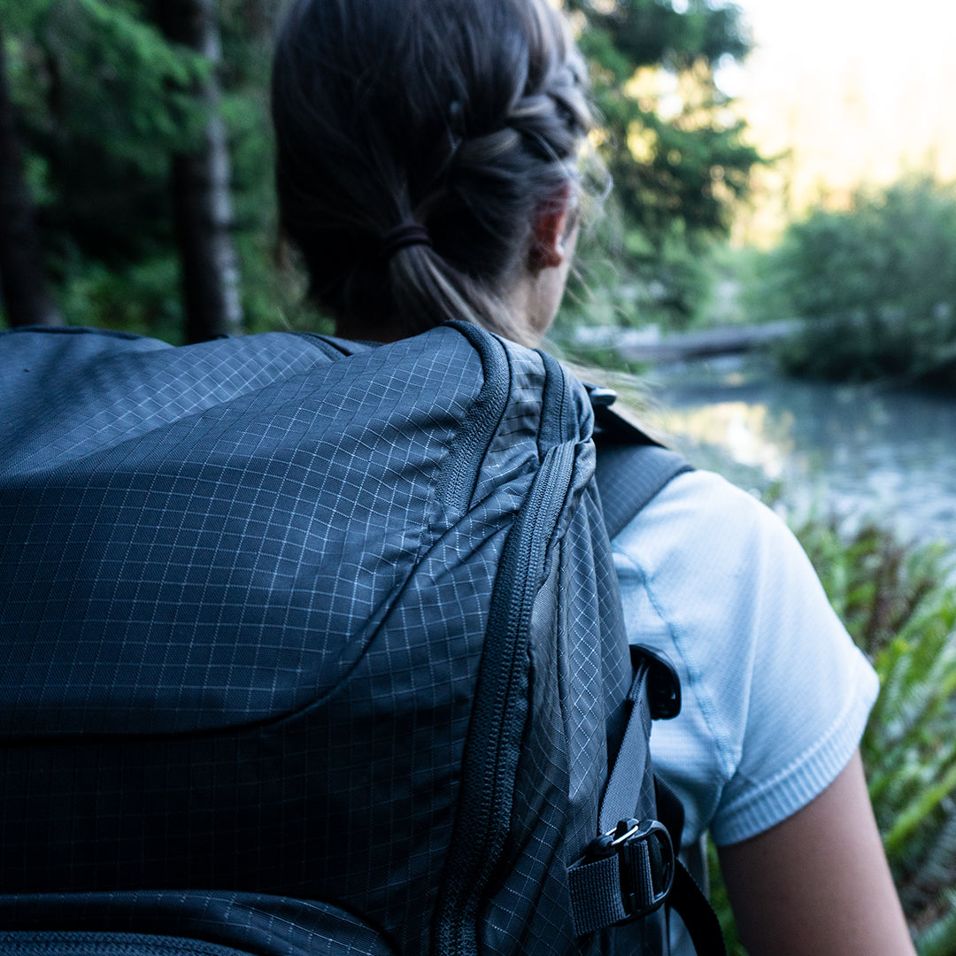 Close-up rear view of a person wearing the Outdoor Vitals KotaUL Ultralight Travel & Adventure Backpack, highlighting the durable grid-patterned ripstop fabric texture against a blurred outdoor forest and river backdrop.