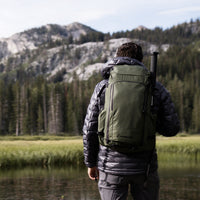 Back view of a hiker wearing a puffy jacket and the Outdoor Vitals KotaUL Ultralight Travel & Adventure Backpack with a trekking pole attached, standing near a mountain lake with pine trees and rocky peaks in the background.