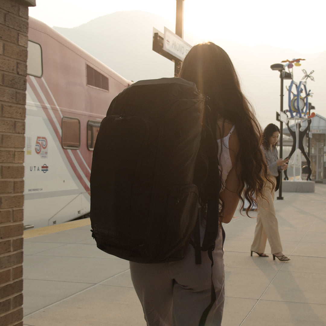 Back view of a traveler wearing the Outdoor Vitals KotaUL Ultralight Travel & Adventure Backpack while walking on a train station platform next to a commuter train.